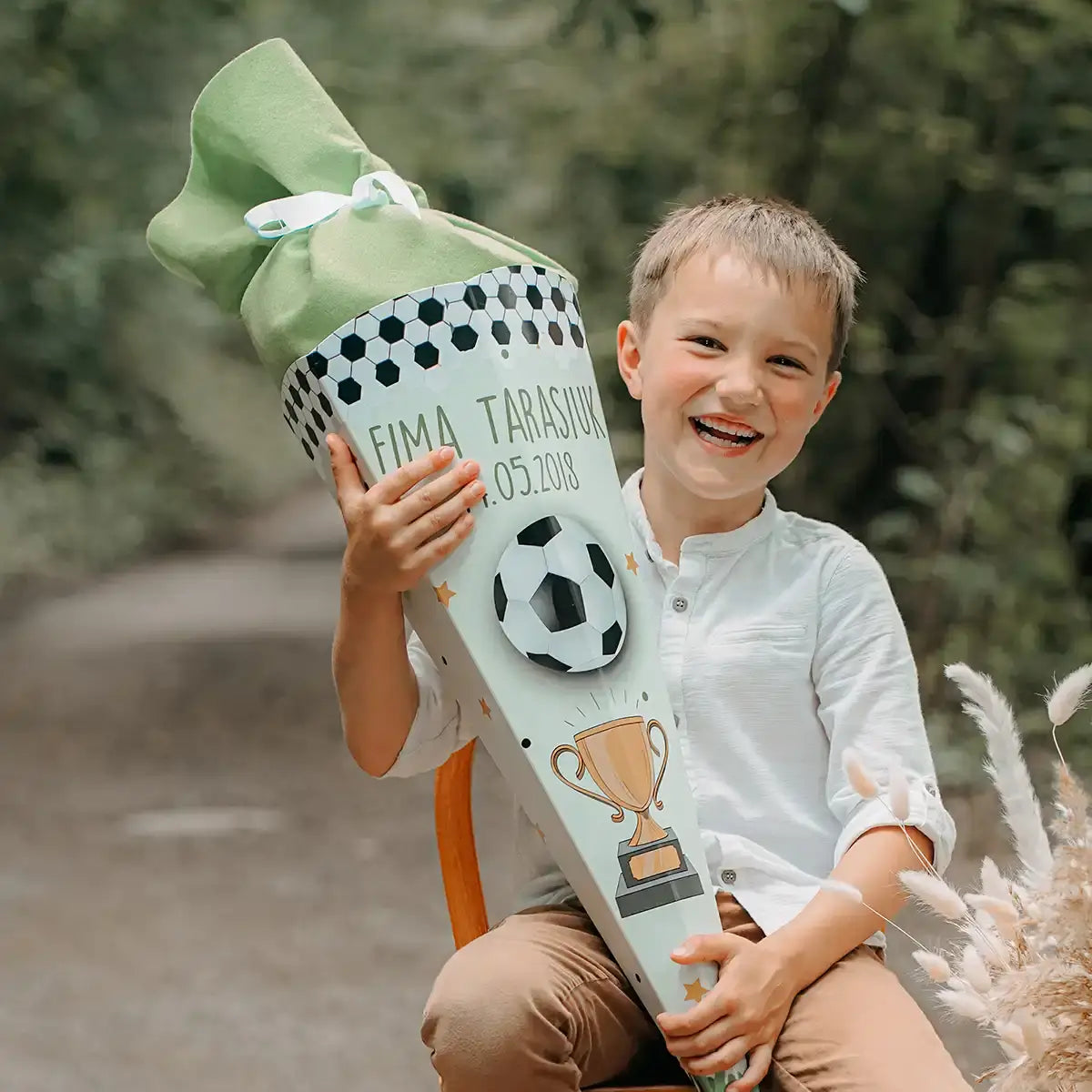 Strahlender Junge mit Fußball-Schultüte zum Schulanfang - glücklicher Erstklässler präsentiert seine personalisierte Schultüte mit Sportmotiven