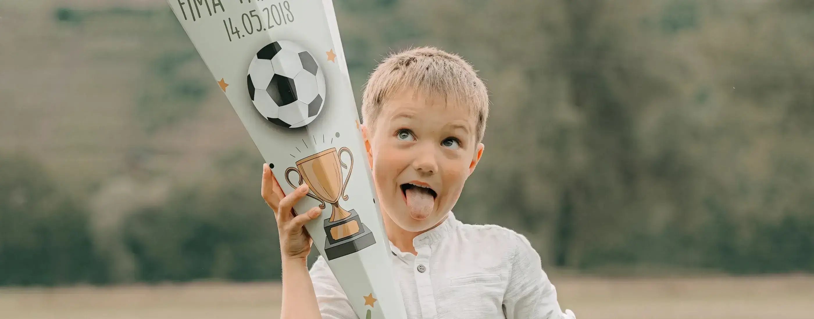 Fröhlicher Junge mit herausgestreckter Zunge hält stolz seine Fußball-Schultüte zur Einschulung mit Pokal und Fußball-Motiv, personalisiert mit Datum 14.05.2019