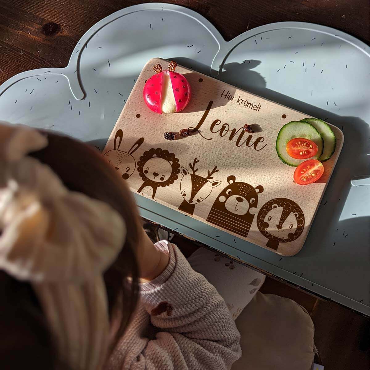 Personalisiertes Kinderfrühstücksbrettchen aus Holz mit Namen 'Leonie', niedlichen Waldtier-Gravuren und dem Spruch 'Hier krümelt'. Auf dem Brettchen liegen ein Marienkäfer-Snack, Gurken- und Tomatenscheiben. Ideal für kinderfreundliches Frühstück auf einer wolkenförmigen blauen Unterlage