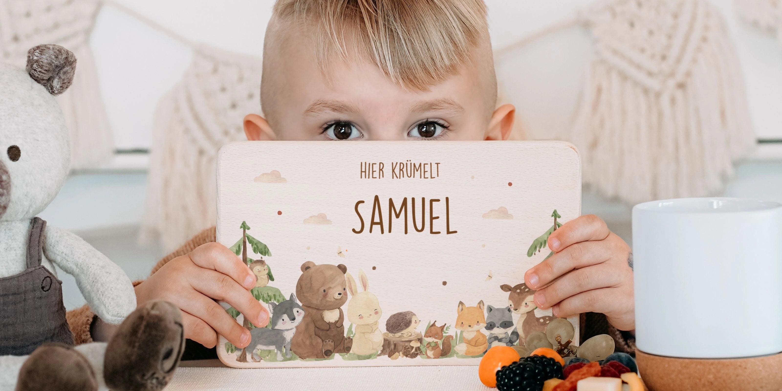 Kleiner Junge hält personalisiertes Frühstücksbrettchen mit Waldtiere-Motiv und Gravur 'Hier krümelt Samuel', umgeben von Kuscheltieren, Obst und einem Becher