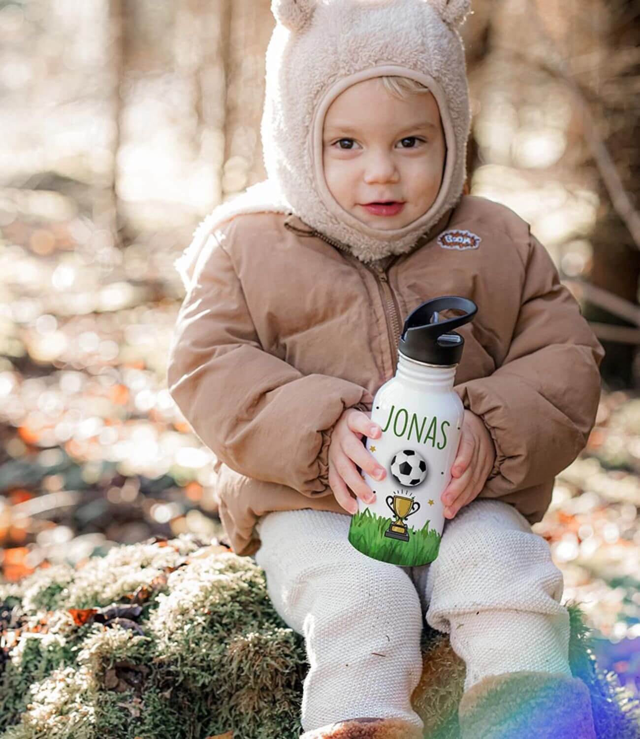 Kleinkind in beiger Winterjacke mit personalisierter Trinkflasche im Wald - lächelnder Junge mit Mütze hält seine Wasserflasche mit aufgedrucktem Namen 'Jonas' und Fußballmotiv in den Händen